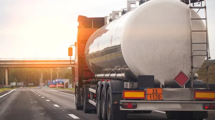Tanker truck with hazardous materials signage driving on highway at sunset, metallic tank gleaming in golden light.