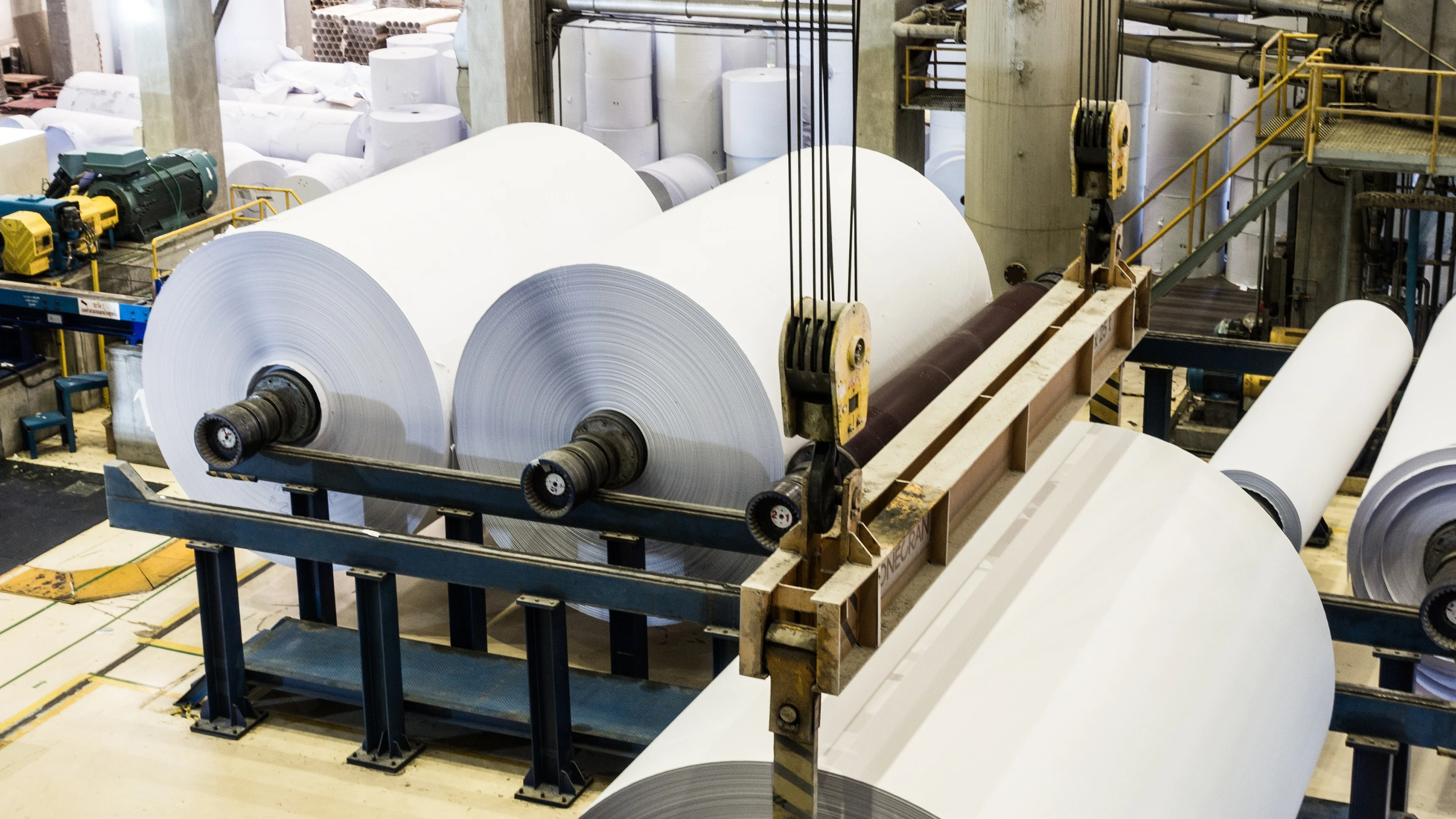 Large white paper rolls on industrial machinery in a paper manufacturing facility with conveyor systems.